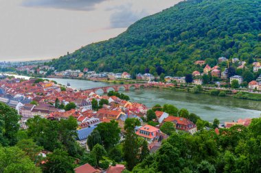 Dramatic aerial view of Heidelberg's old town, featuring colorful buildings along the Neckar River with lush green hills in the background under a cloudy sky.
