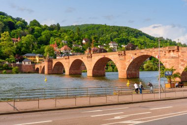 Heidelberg, Almanya - 8 Haziran 2025: Neckar Nehri boyunca uzanan kemerli taş köprü, tepelerde canlı yeşillikler ve arka planda ilginç binalar, sakin bir atmosfer yakalıyor.