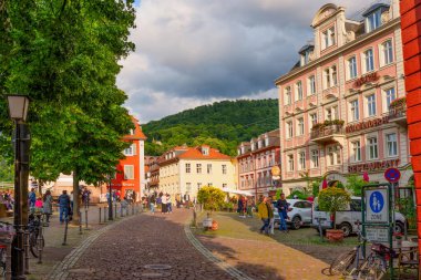 Heidelberg, Almanya - 8 Haziran 2025: Heidelberg 'de tarihi binalar, yemyeşil alanlar ve manzaranın tadını çıkaran yayaların yer aldığı canlı sokak manzarası.