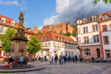 Heidelberg, Almanya - 8 Haziran 2025: Tarihi binalar ve hareketli ziyaretçilerle çevrili büyüleyici Kornmarkt Madonna çeşmesi, Heidelberg Kalesi 'nin tepesinde parçalı bulutlu bir gökyüzünün altında görünür.