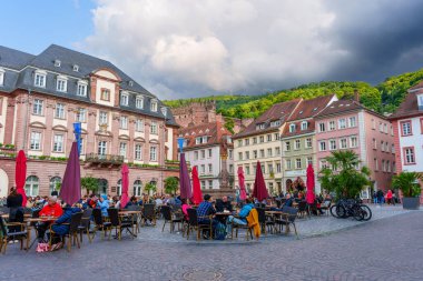 Heidelberg, Almanya - 8 Haziran 2025: Heidelberg 'in tarihi meydanında canlı bir sahne, açık hava kafeleri, sakin yemeklerden hoşlanan müşteriler ve dramatik bir gökyüzü altında klasik Alman mimarisi.