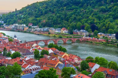 Heidelberg, Almanya - 8 Haziran 2025: Eski Köprü 'nün panoramik manzarası, renkli çatılar ve yumuşak günbatımı parlaklığında yemyeşil yamaçlar.