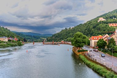Heidelberg, Almanya - 8 Haziran 2025: Huzurlu Neckar Nehri, tarihi köprü ve bulutlu bir gökyüzü altındaki yemyeşil tepelerin yer aldığı Heidelberg 'in dramatik manzarası.