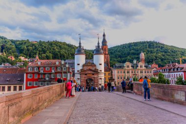 Heidelberg, Almanya - 8 Haziran 2025: Heidelberg 'deki Eski Köprü manzarası, ikonik kuleler, tarihi binalar ve dramatik bir gökyüzü altında yemyeşil bir tepe..