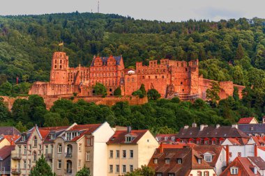 Heidelberg, Almanya - 8 Haziran 2025: Heidelberg Kalesi 'nin panoramik manzarası, günbatımı parlaklığıyla aydınlanan, tarihi mimarinin bir karışımını sergileyen, ön planda tuhaf binalar bulunan bir tepeye tünemiştir..
