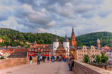 Heidelberg, Almanya - 8 Haziran 2025: Heidelberg 'deki Eski Köprü' de turistlerin hareketli manzarası, dramatik bir gökyüzü altında resimli mimari ve yemyeşil tepeleri gözler önüne seriyor.