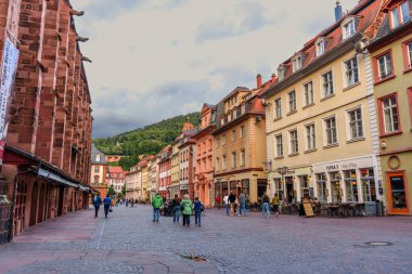 Heidelberg, Almanya - 8 Haziran 2025: Heidelberg 'de renkli binalar, kaldırımlar, yayalar ve bulutlu bir gökyüzü altında yeşil tepelerin yer aldığı canlı sokak manzarası.
