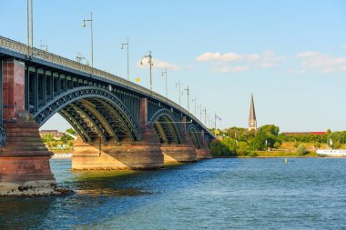 Mainz, Almanya - 19 Haziran 2025: Ren Nehri boyunca uzanan tarihi bir köprünün manzarası, Mainz 'de berrak mavi bir gökyüzünün altında görkemli kemler ve önemli bir kilise kulesi yer alıyor..