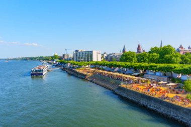 Mainz, Almanya - 19 Haziran 2025: Mainz 'de güneş banyosu yapanlar, Ren Nehri' ndeki kayıklar ve yemyeşil ağaçların yer aldığı canlı nehir kıyısı sahnesi.