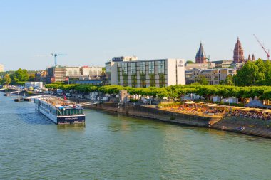 Mainz, Almanya - 19 Haziran 2025: Mainz 'deki Ren Nehri' nin panoramik manzarası, parlak güneş ışığı altındaki bir nehir teknesi, canlı bir rıhtım ve tarihi binalar.