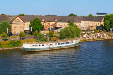 Mainz, Almanya - 19 Haziran 2025: Berrak mavi gökyüzü altında yemyeşil ve tarihi binalarla çevrili Mainz 'deki nehir kıyısında terk edilmiş tekne.