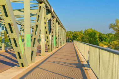 Mainz, Almanya - 19 Haziran 2025: Açık mavi gökyüzü altındaki canlı yeşilliklerle çevrili nehir boyunca uzanan çelik köprü yolu.