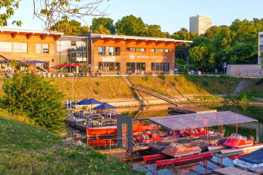 Mainz, Almanya - 19 Haziran 2025: Mainz Kürek Kulübü 'ne yanaşan renkli tekneler, yemyeşil ağaçların ve modern yapının arkasında güneş batıyor..