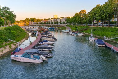 Mainz, Almanya - 19 Haziran 2025: Günbatımında sakin marina sahnesi, ağaçlar ve konut binalarıyla çevrili güzel bir su yolu ile demirlenmiş çeşitli teknelerin yer aldığı.