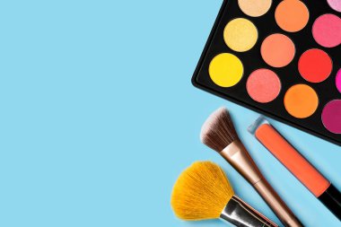 Parlak renkli sarı, kırmızı, pembe, turuncu göz farı, turuncu sıvı lipgloss ve pastel mavi arka plan ile stüdyo ışık yukarıdan ateş köşesinde düzenlenmiş iki makyaj fırçaları siyah plastik palet