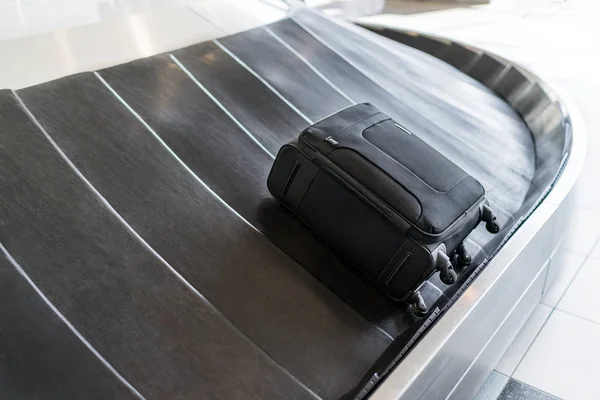 A small-sized black luggage bag is laying on a rotating black rubber airport baggage claim carousel (conveyor belt) with a shiny steel body frame in natural daylight in the morning