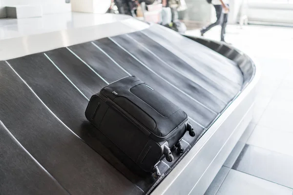 A small-sized black luggage bag is laying on a rotating black rubber airport baggage claim carousel (conveyor belt) with a shiny steel body frame in natural daylight in the morning