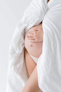 Bebeğin doğumu beklentisiyle mutlu genç anne. Hamile bir kız beyaz bir çarşaf ile kaplı çıplak karnını çırparak duruyor. Beyaz arka planda.