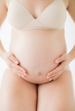 Bebeğin doğumu beklentisiyle mutlu genç anne. Hamile bir kız çıplak karnını çırparak oturuyor. Beyaz arka planda.