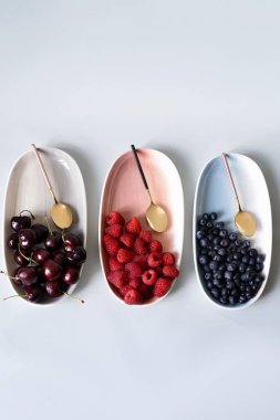 Yaz meyveleri yaban mersini, ahududu ve kiraz pembe, mavi ve gri renklerin yeni zarif tabaklar üzerinde. İzole beyaz arka plan üzerinde yukarıdan yeni lüks Altın çatal görünümü. Üstte görüntü.