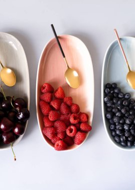 Yaz meyveleri yaban mersini, ahududu ve kiraz pembe, mavi ve gri renklerin yeni zarif tabaklar üzerinde. İzole beyaz arka plan üzerinde yukarıdan yeni lüks Altın çatal görünümü. Üstte görüntü.