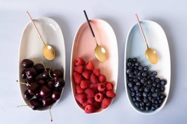 Yaz meyveleri yaban mersini, ahududu ve kiraz pembe, mavi ve gri renklerin yeni zarif tabaklar üzerinde. İzole beyaz arka plan üzerinde yukarıdan yeni lüks Altın çatal görünümü. Üstte görüntü.