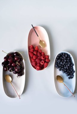 Yaz meyveleri yaban mersini, ahududu ve kiraz pembe, mavi ve gri renklerin yeni zarif tabaklar üzerinde. İzole beyaz arka plan üzerinde yukarıdan yeni lüks Altın çatal görünümü. Üstte görüntü.