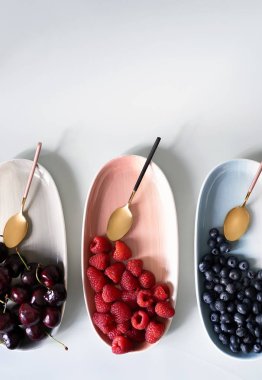 Yaz meyveleri yaban mersini, ahududu ve kiraz pembe, mavi ve gri renklerin yeni zarif tabaklar üzerinde. İzole beyaz arka plan üzerinde yukarıdan yeni lüks Altın çatal görünümü. Üstte görüntü.