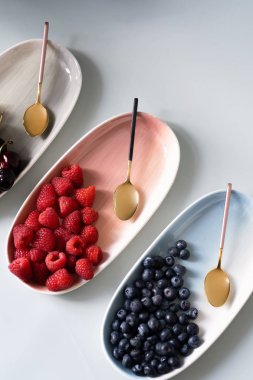 Yaz meyveleri yaban mersini, ahududu ve kiraz pembe, mavi ve gri renklerin yeni zarif tabaklar üzerinde. İzole beyaz arka plan üzerinde yukarıdan yeni lüks Altın çatal görünümü. Üstte görüntü.