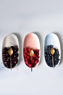 Yaz meyveleri yaban mersini, ahududu ve kiraz pembe, mavi ve gri renklerin yeni zarif tabaklar üzerinde. İzole beyaz arka plan üzerinde yukarıdan yeni lüks Altın çatal görünümü. Üstte görüntü.