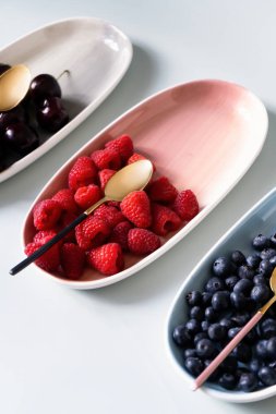 Yaz meyveleri yaban mersini, ahududu ve kiraz pembe, mavi ve gri renklerin yeni zarif tabaklar üzerinde. İzole beyaz arka plan üzerinde yukarıdan yeni lüks Altın çatal görünümü. Üstte görüntü.