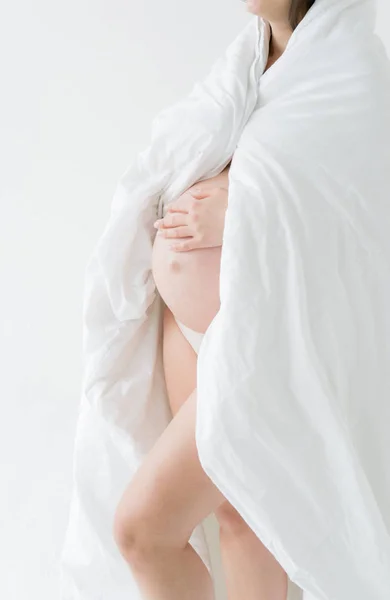 赤ちゃんの誕生を見越して幸せな若い母親 妊娠中の女の子は白いシートで覆われた裸の腹を抱いて立っています 白い背景に — ストック写真