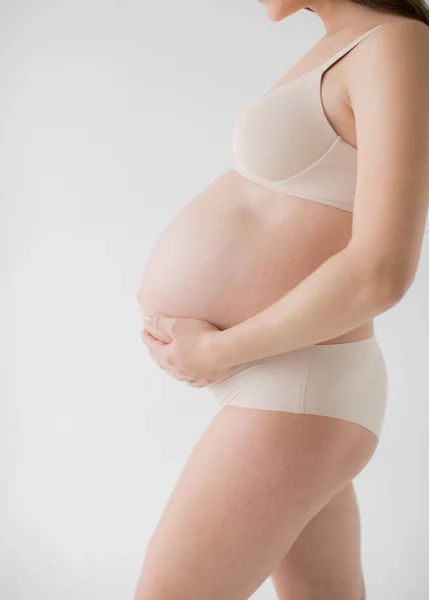 Bebeğin doğumu beklentisiyle mutlu genç anne. Hamile bir kız elleriyle çıplak karnını çırpıyor. Beyaz arka planda.