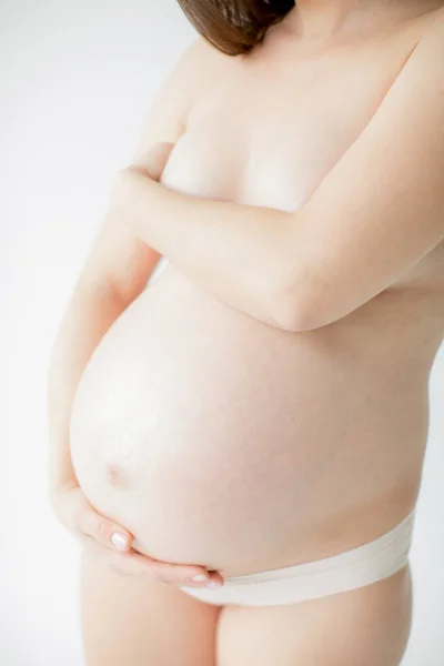 Bebeğin doğumu beklentisiyle mutlu genç anne. Hamile bir kız elleriyle çıplak karnını çırpıyor. Beyaz arka planda.