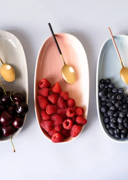 Yaz meyveleri yaban mersini, ahududu ve kiraz pembe, mavi ve gri renklerin yeni zarif tabaklar üzerinde. İzole beyaz arka plan üzerinde yukarıdan yeni lüks Altın çatal görünümü. Üstte görüntü.