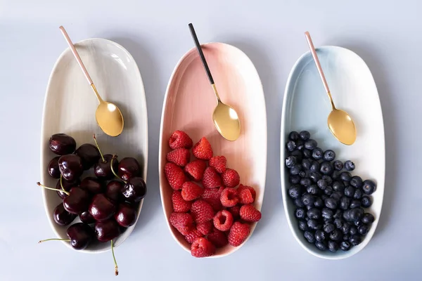 Yaz meyveleri yaban mersini, ahududu ve kiraz pembe, mavi ve gri renklerin yeni zarif tabaklar üzerinde. İzole beyaz arka plan üzerinde yukarıdan yeni lüks Altın çatal görünümü. Üstte görüntü.