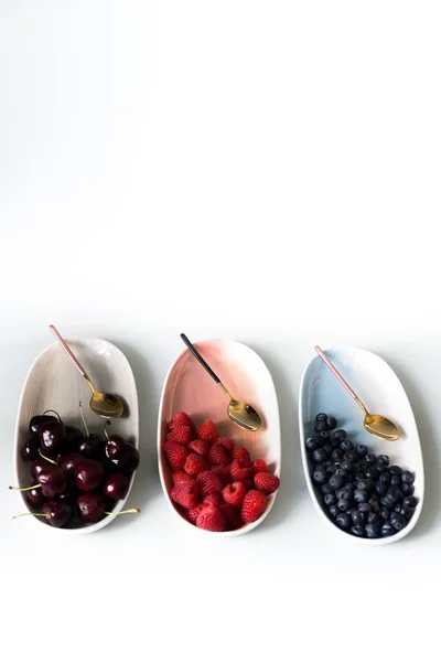 Yaz meyveleri yaban mersini, ahududu ve kiraz pembe, mavi ve gri renklerin yeni zarif tabaklar üzerinde. İzole beyaz arka plan üzerinde yukarıdan yeni lüks Altın çatal görünümü. Üstte görüntü.
