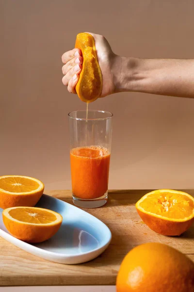 Dişi el, taze soğuk portakalı bardağa sıkıştırır. Tahta bir tahta ve mavi bir tabak ta dilimlenmiş narenciye. Kahvaltıda portakal suyu, sağlık için vitaminler.