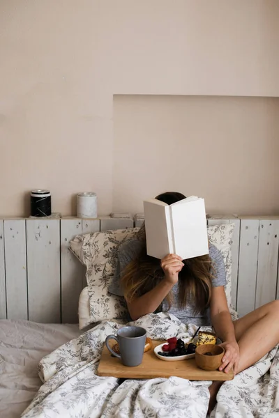 Lazy morning in warm bed. The girl has breakfast in the morning in her room. Beautiful and cute girl reading book in the winter a cozy atmosphere. Eating fresh berries, sweet piece of cake, hot tea.