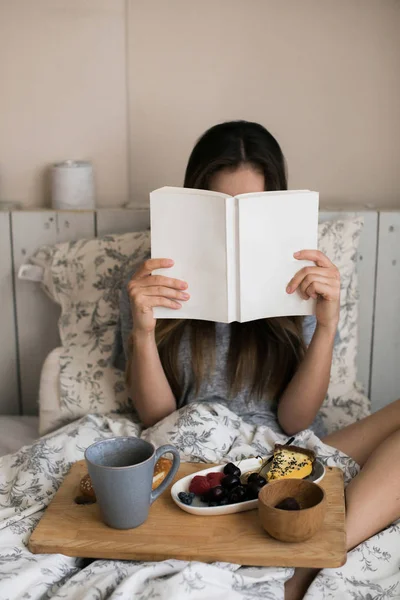 Lazy morning in warm bed. The girl has breakfast in the morning in her room. Beautiful and cute girl reading book in the winter a cozy atmosphere. Eating fresh berries, sweet piece of cake, hot tea.