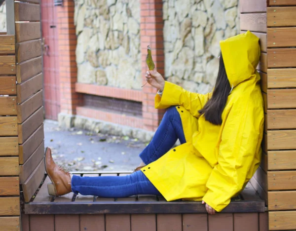 girl in a raincoat holding fallen leaves in the fall. A man sits in a raincoat on a wooden bench.