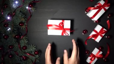 Xmas arka plan siyah ahşap çam dalları ile dekore edilmiştir. Baubles kırmızı konfeti ile süslenmiş ve parıldayan ışıklar çam. Kırmızı kurdele ile beyaz hediyeler. Bir çocuk onu masaya bir hediye verdi. Üstten görünüm, tebrik mesajı alan. 4 k çözünürlükte filme