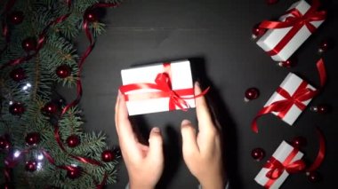 Xmas arka plan siyah ahşap çam dalları ile dekore edilmiştir. Baubles kırmızı konfeti ile süslenmiş ve parıldayan ışıklar çam. Kırmızı kurdele ile beyaz hediyeler. Bir çocuk bir hediye masanın üzerine koyar. Üstten görünüm, tebrik mesajı alan. 4 k çözünürlükte filme