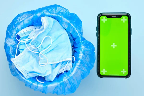 Disposable used surgical mask in a trash bin and smartphone with chroma key on a blue background. Application of smart quarantine concept
