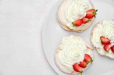 Beyaz arka planda beyaz bir tabakta, üzerinde taze çilek bulunan üç Pavlova kremalı kekin üst görüntüsü. Ev yapımı pasta..