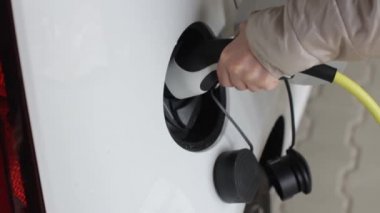 Kadın elektrikli beyaz bir arabanın şarj prizini çıkarmış. Çevre dostu ve sıfır emisyonlu araba.