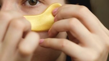 Genç bir kadın gözlerinin altında altın kolajen hidrojel göz bandı kullanıyordu. Kırışıklıkların ortadan kalkması. Evde cilt bakımı. Kozmetoloji prosedürü. 