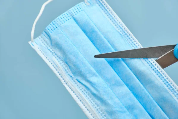 Scissors cutting a surgical face mask against coronavirus disease on a blue background. Cut mask before before disposing — Stock Photo, Image