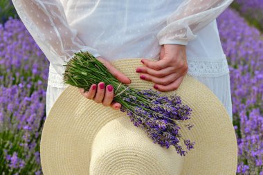 Beyaz açık renk elbiseli genç bir kadın arkasında bir şapka ve mor bir demet lavanta tutuyordu.. 