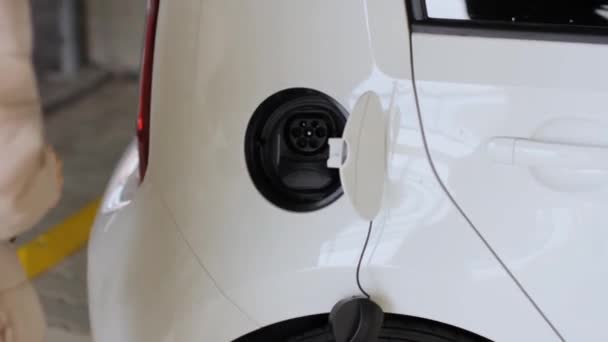 Das Anbringen eines Stromkabels an einem Elektroauto durch eine Frau in einem Licht schließt sich. Laden von Elektrofahrzeugen. — Stockvideo